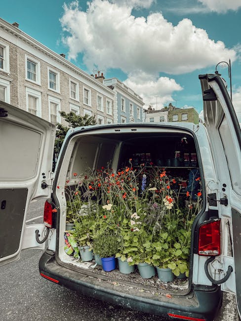 The open rear of a white van filled with potted plants and fresh flowers, including red and white blooms with green foliage, arranged in blue and gray pots. The flowers appear vibrant and freshly pick