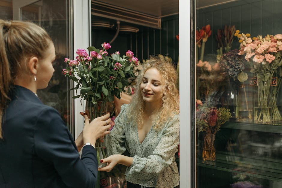 A young woman with curly blonde hair happily receives a bouquet of fresh pink roses with green foliage from a floral specialist at North Kensington Florist, inside a refrigerated display case. The flo