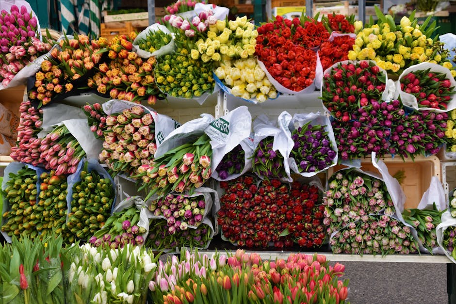 A vibrant display of freshly cut flowers arranged in bouquets and bundles at a flower shop, featuring a variety of blooms including tulips in shades of pink, white, red, purple, and green. The flowers