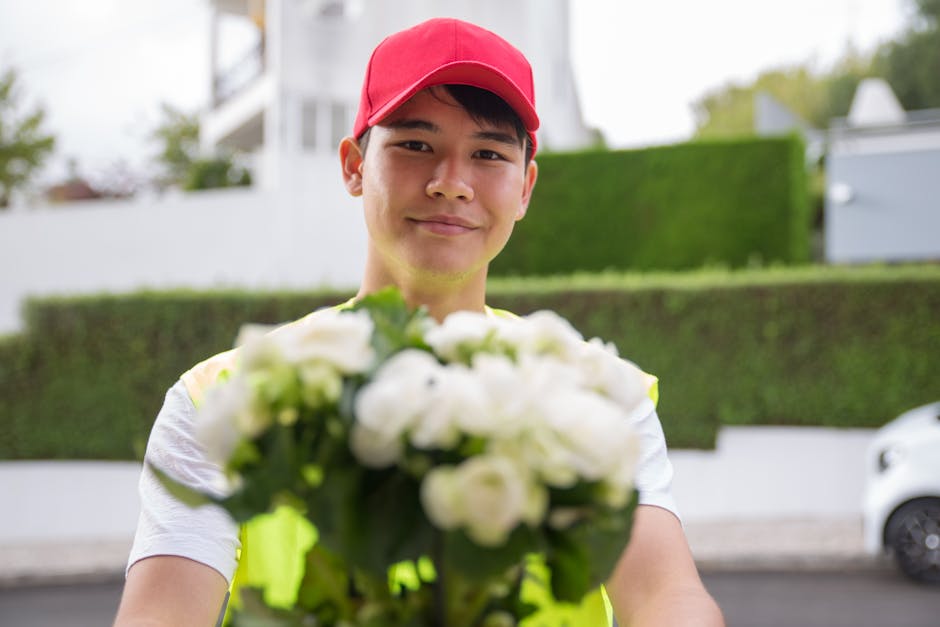 A delivery person wearing a red cap and a reflective safety vest is holding a bouquet of fresh white flowers, including roses and possibly tulips, wrapped in clear plastic with some greenery. The bouq