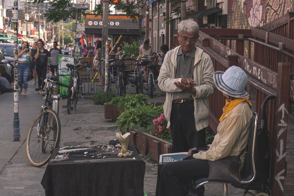 A street scene in North Kensington featuring an elderly man with gray hair, wearing glasses, a light gray jacket, and dark pants, standing outdoors. He is handing over a small item to a seated woman w