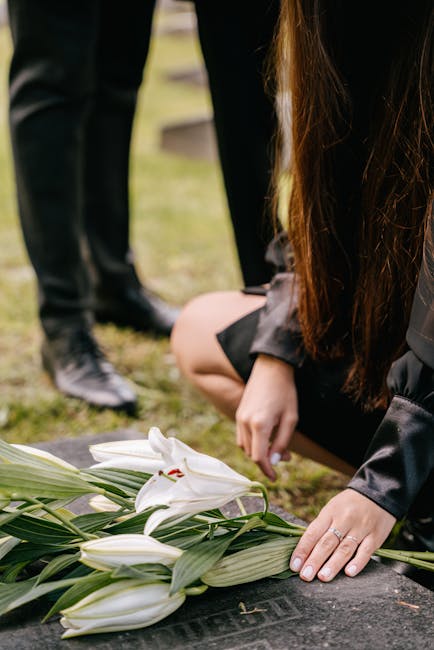 A person kneeling on the ground, arranging a floral tribute consisting of white lilies with large, open petals and green stems with long, pointed leaves. The flowers are resting on a dark stone surfac