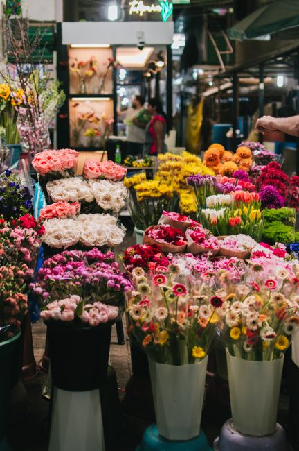 A vibrant flower stall at an indoor market featuring an extensive display of fresh flowers arranged in bouquets and wrapped in various colors. The flowers include soft pink and white roses, cheerful y
