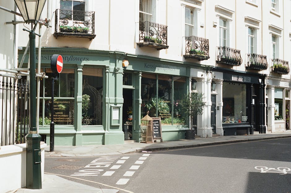 A view of a floral shop on a street corner with large glass windows displaying fresh flowers inside. The shop has a light green facade with decorative molding and a sign that reads 'Kensington Flowers