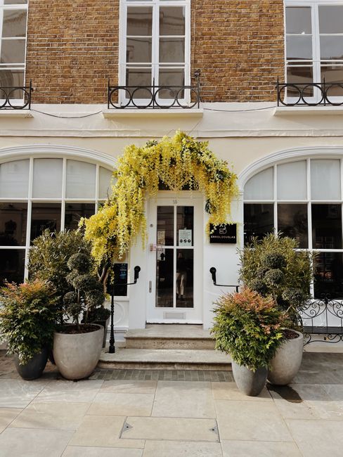 The image depicts the entrance to a florist shop, with a white door framed by two large, curved windows. Above the entrance, a lush floral display features cascading yellow wisteria and white flowers,