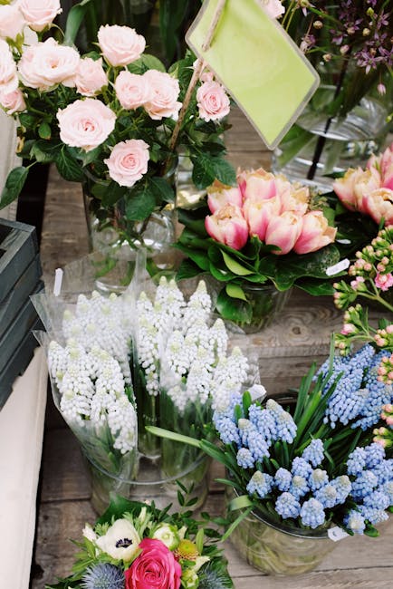 A display of fresh floral arrangements on a rustic wooden surface, featuring a tall bouquet of pale pink roses with dark green foliage in a clear glass vase, alongside a round arrangement of pink and 