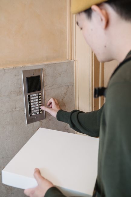 A person wearing a yellow beanie and dark green jacket is using a key pad for access control, pressing a button with their right hand. They are holding an empty white box with their left hand, prepari