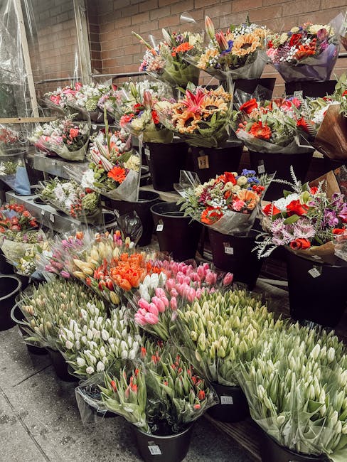 A display of numerous fresh flower bouquets and bunches arranged in black buckets and wrapped in floral paper, showcasing a variety of vibrant colors including pink, orange, red, purple, and white. Th