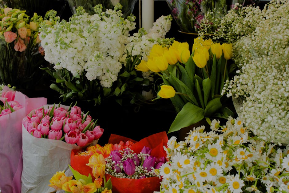 A vibrant display of fresh floral arrangements featuring a variety of flowers including yellow tulips with elongated green stems and broad leaves, white daisies with bright yellow centers, pink tulips